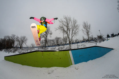 Snowboarder performing a jump with a Pink Cloud Pro Mini Snowboard 135 on a snowy day, showcasing freestyle skills and vibrant gear.